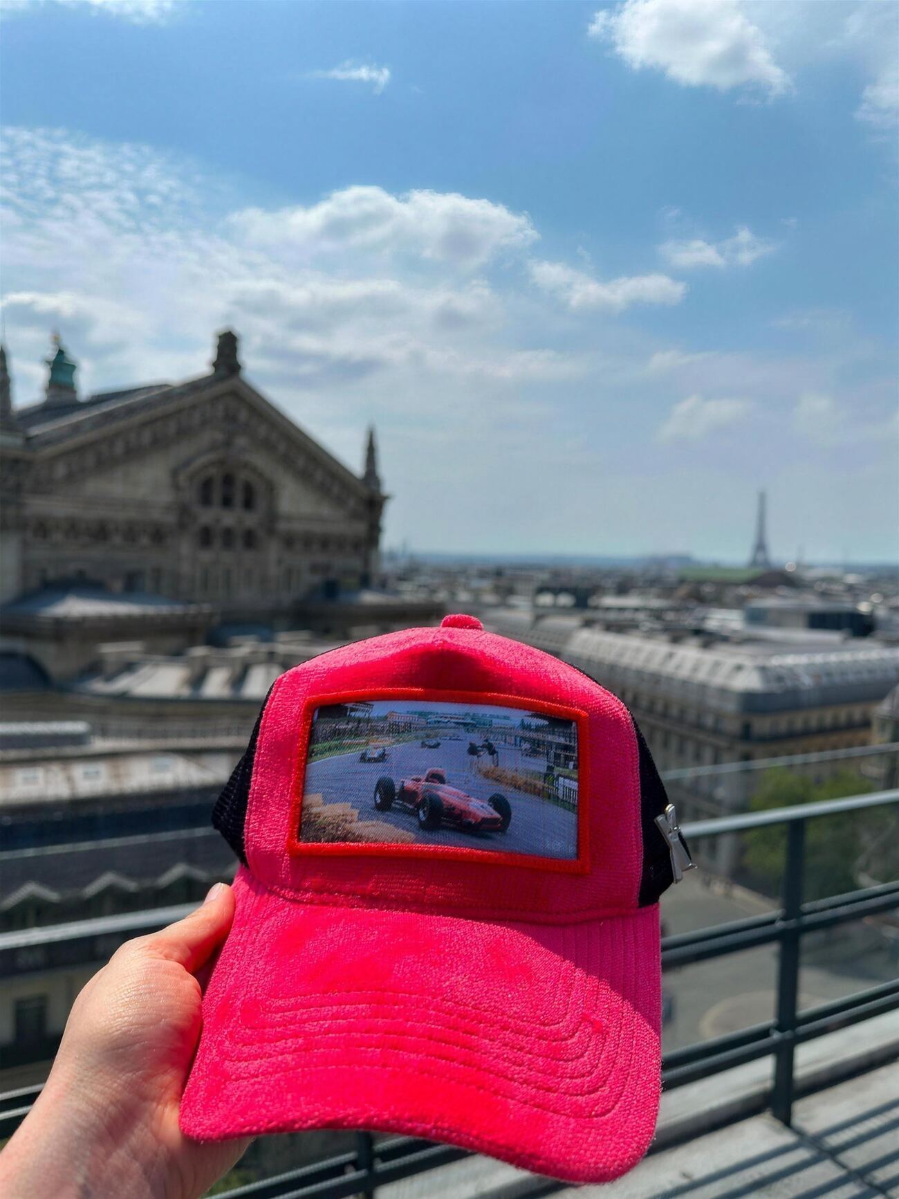 Bright red velvet trucker cap with a vintage grand prix racing patch, held above the Paris rooftops with the Eiffel Tower on the horizon
