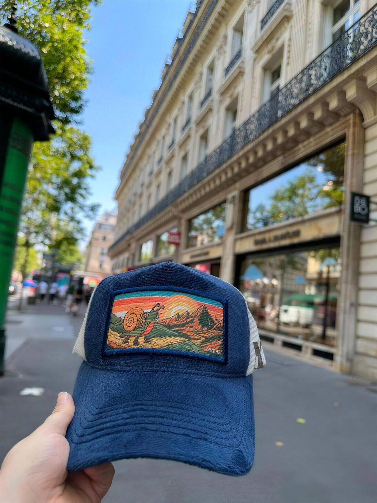 Deep blue velvet trucker cap with a colorful woven patch of a hiking snail before sunrise mountains, on a Haussmann boulevard