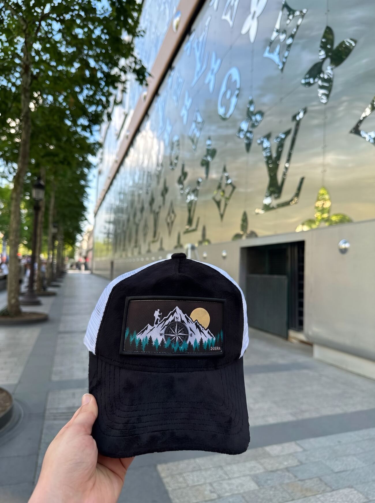 Black and white mesh trucker cap with a woven compass-and-mountains patch, on the Champs-Élysées at dusk