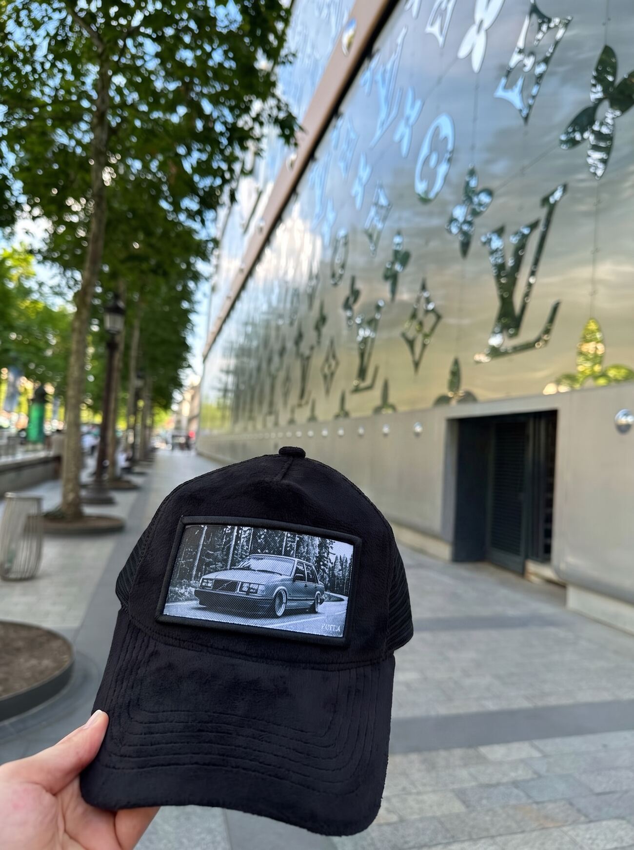 Black velvet trucker cap with a monochrome Volvo 740 woven patch, held against the mirrored monogram facade on the Champs-Élysées
