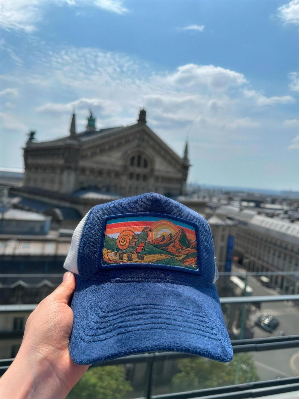 Blue and white mesh trucker cap with a snail and mountains patch on a rooftop terrace facing the Palais Garnier