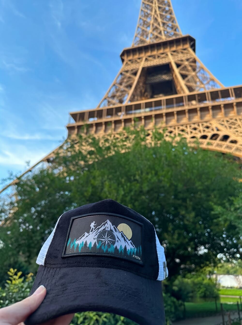 Black and white trucker cap with a mountain compass patch held at the base of the Eiffel Tower