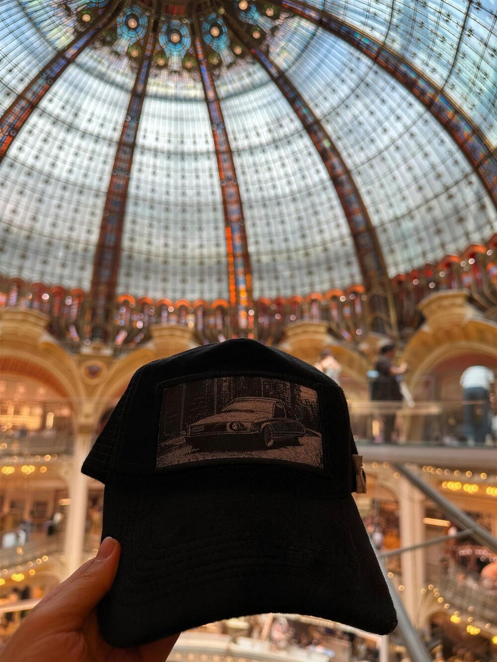Black velvet XVERA trucker cap held up beneath the stained-glass dome of the Galeries Lafayette in Paris